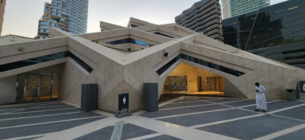 Striking modern architecture of Riyadh's financial district, featuring geometric designs and contrasting sandstone textures, with a figure in traditional attire nearby.
