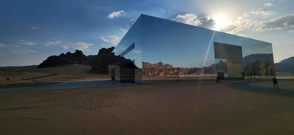 Mirrored Maraya Concert Hall reflecting the desert landscape and sandstone cliffs of AlUla, with visitors exploring the area at sunset.