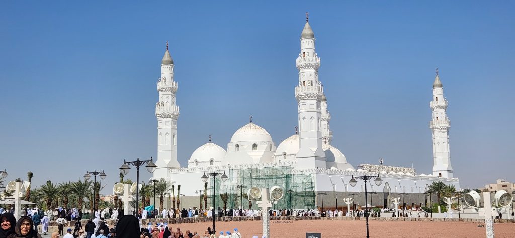 Quba Mosque in Medina, showcasing gleaming white domes and minarets, with pilgrims gathered in the foreground, reflecting the sacred atmosphere of the site.