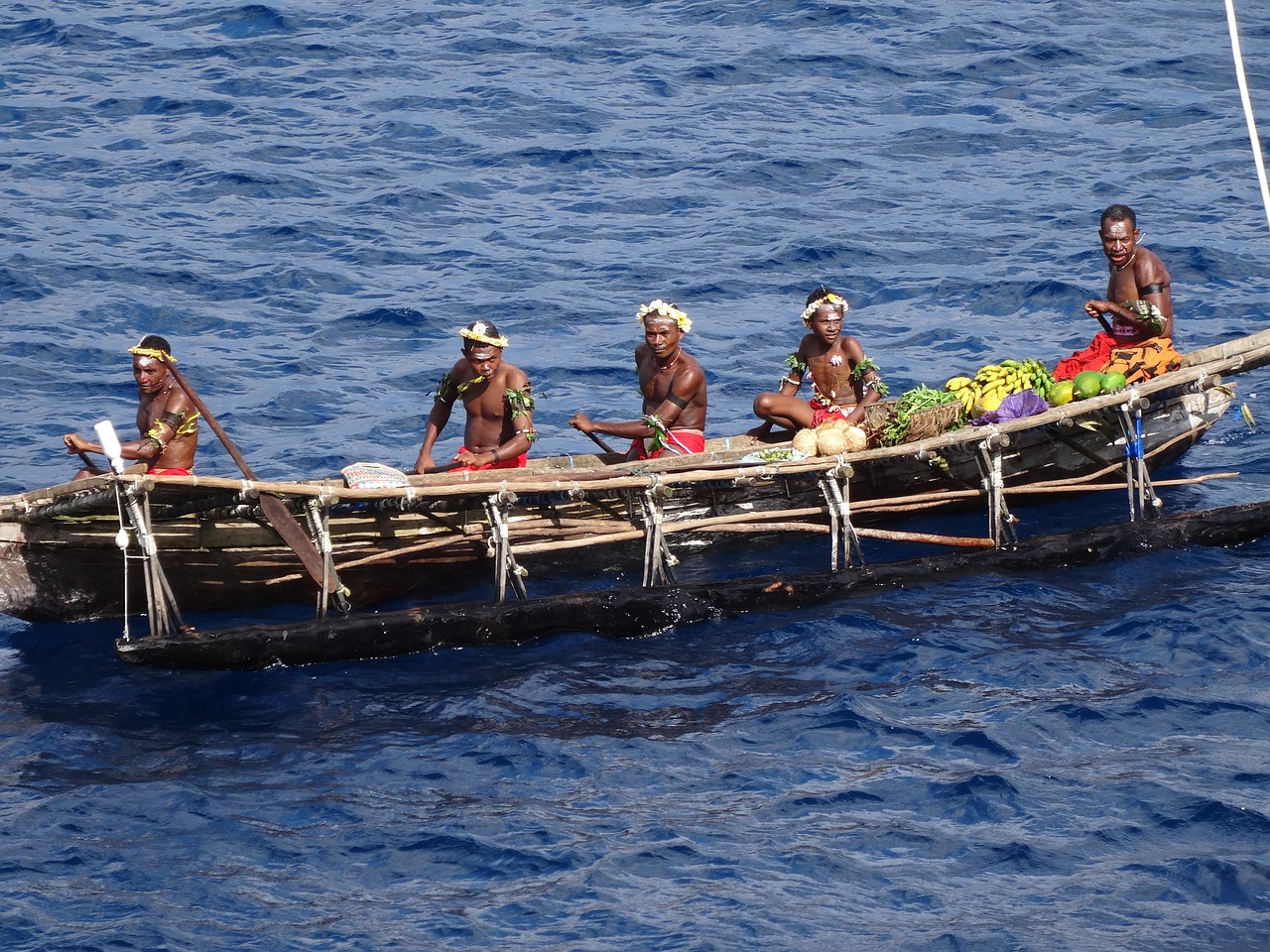 Group of individuals in traditional attire on a canoe, surrounded by tropical fruits, navigating the vibrant blue waters of Papua New Guinea, showcasing local culture and adventure.