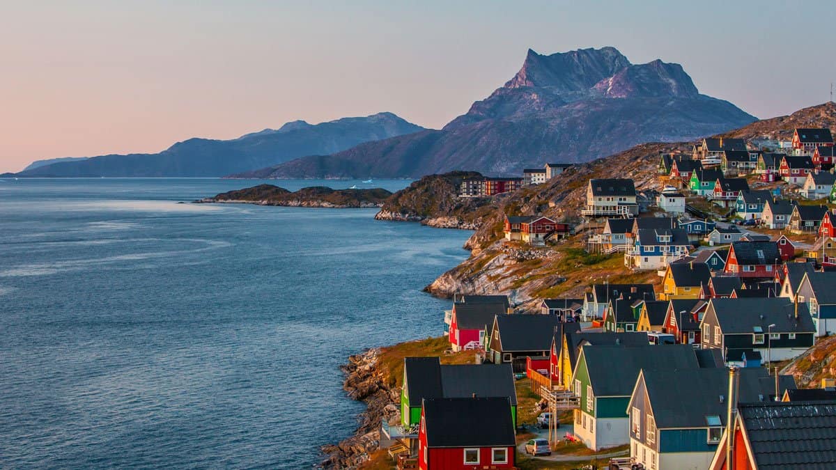 Colorful houses along the coastline of Greenland with mountains in the background at sunset, showcasing the unique Arctic landscape and vibrant local architecture.
