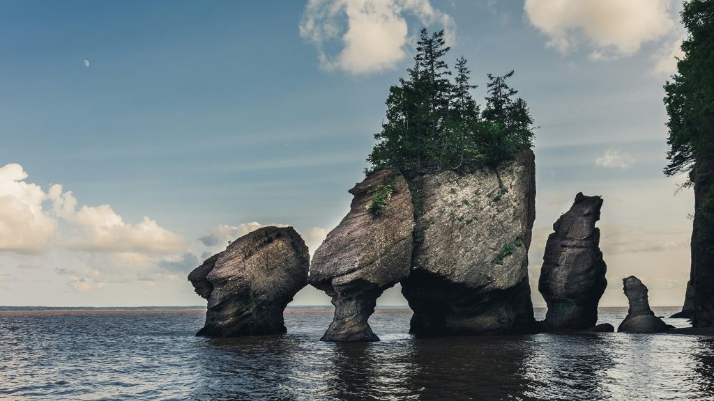 Rock formations with lush greenery on top, surrounded by water under a blue sky with clouds, representing Eastern Canada's natural wonders for travel exploration.
